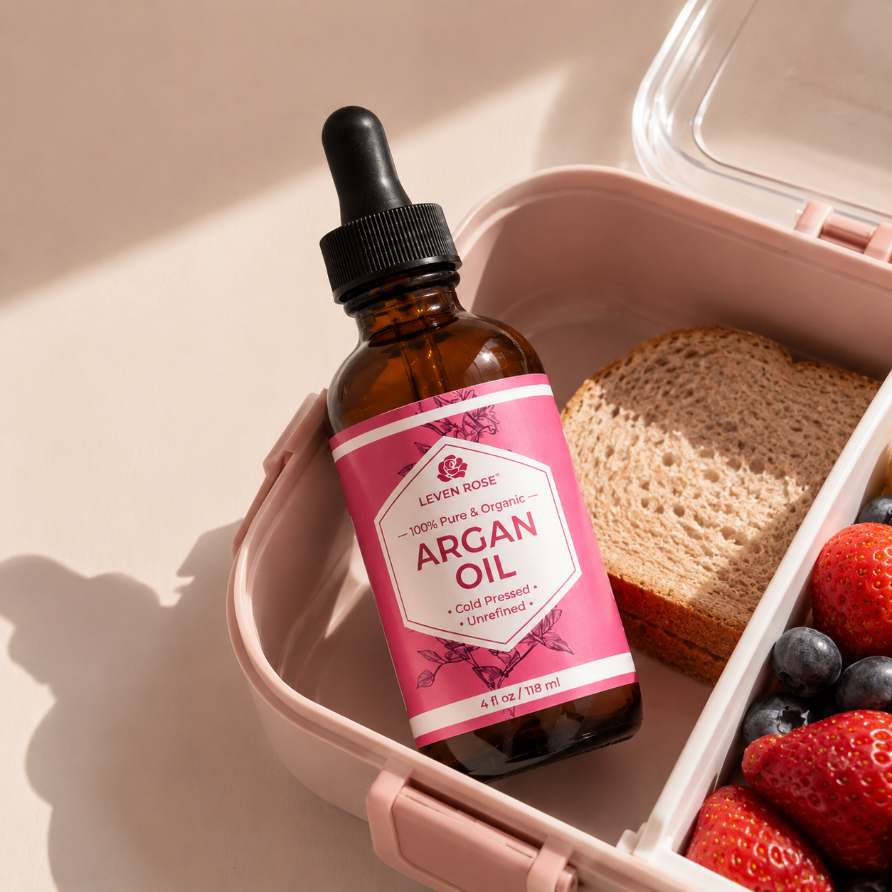 An amber glass dropper bottle of Leven Rose Argan Oil placed inside a pink lunchbox next to a slice of whole wheat bread and fresh berries.