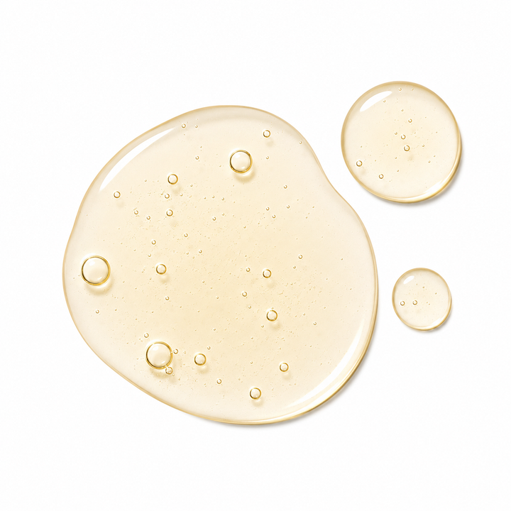 Three translucent droplets of pale golden-yellow cosmetic gel or serum on a clean white background, with the largest droplet spread in an irregular oval shape and two smaller droplets to the upper right.