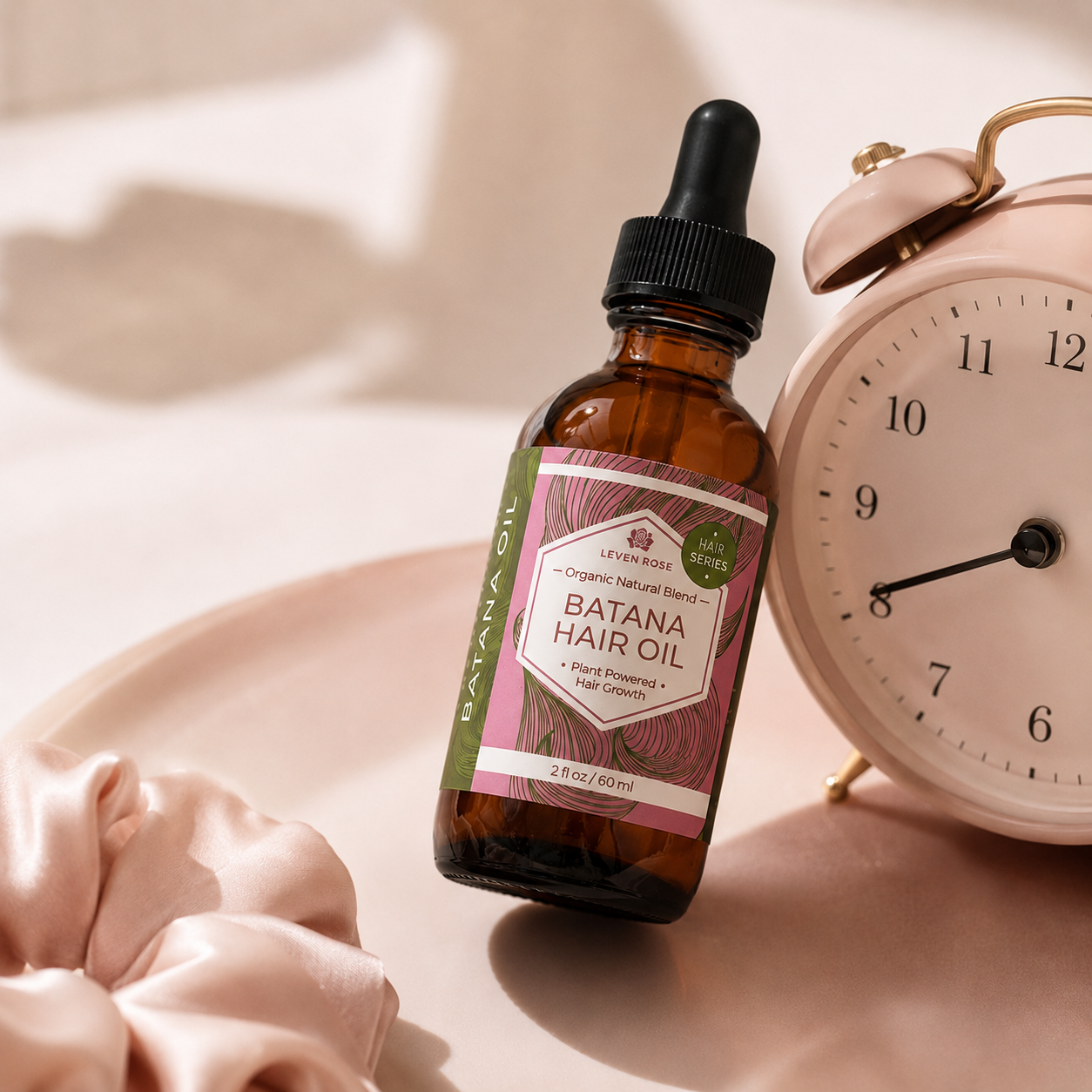 A bottle of Levenwood Botanicals Batana Hair Oil in an amber glass dropper bottle, styled on a beige surface next to a pink alarm clock and a satin scrunchie.