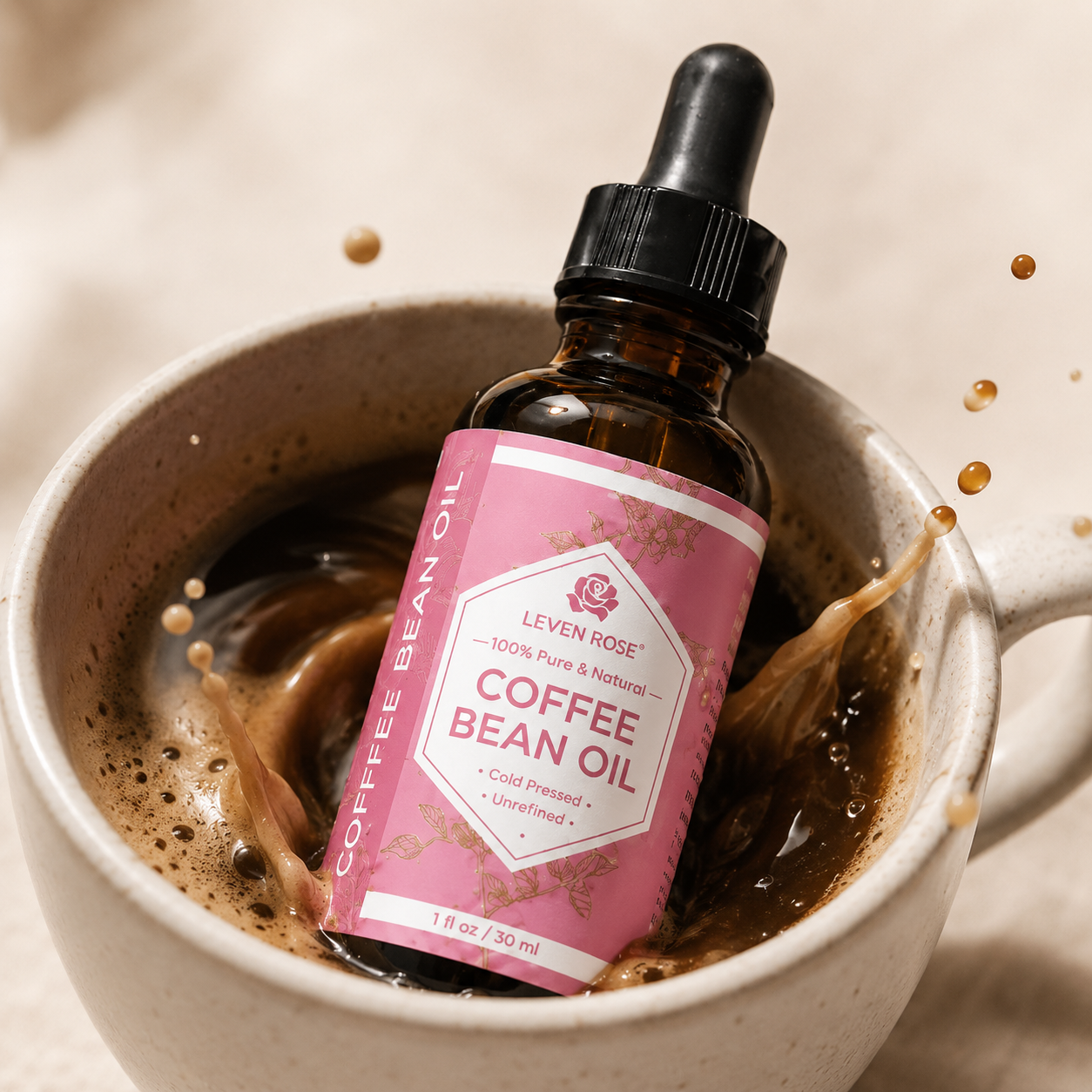 A bottle of Leven Rose Coffee Bean Oil with a pink label and black dropper cap, resting in a ceramic mug filled with coffee and splashing liquid, on a warm beige background.