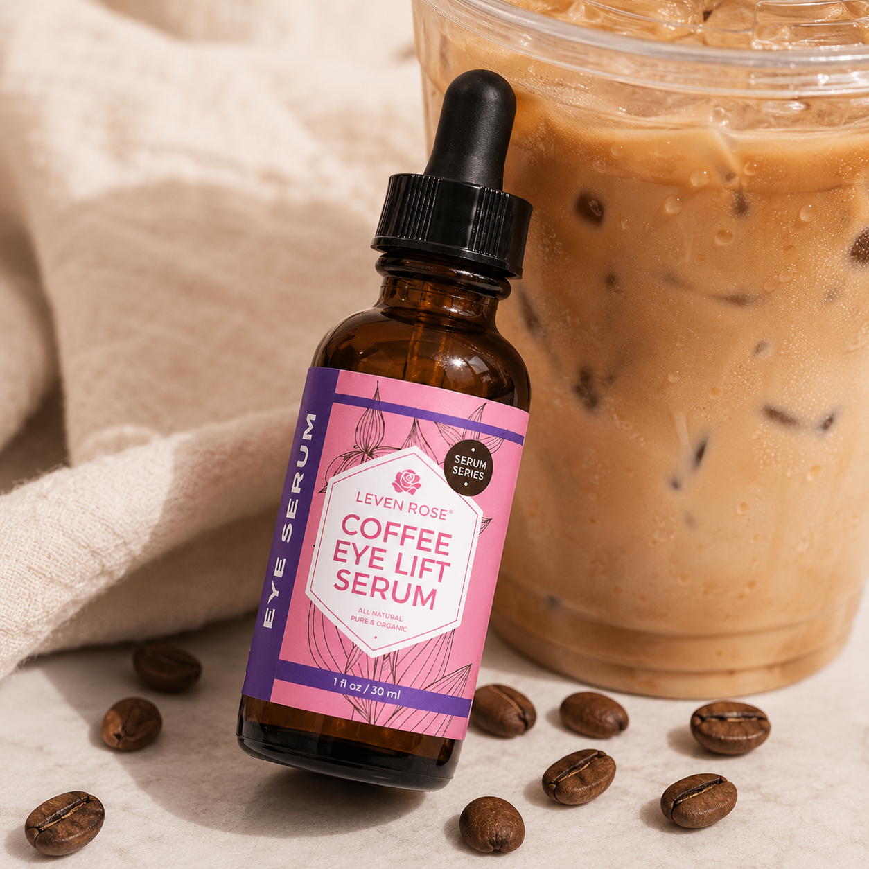 A bottle of Leven Rose Coffee Eye Lift Serum in an amber glass dropper bottle with a pink and purple label, styled next to an iced coffee cup with scattered coffee beans on a neutral surface.