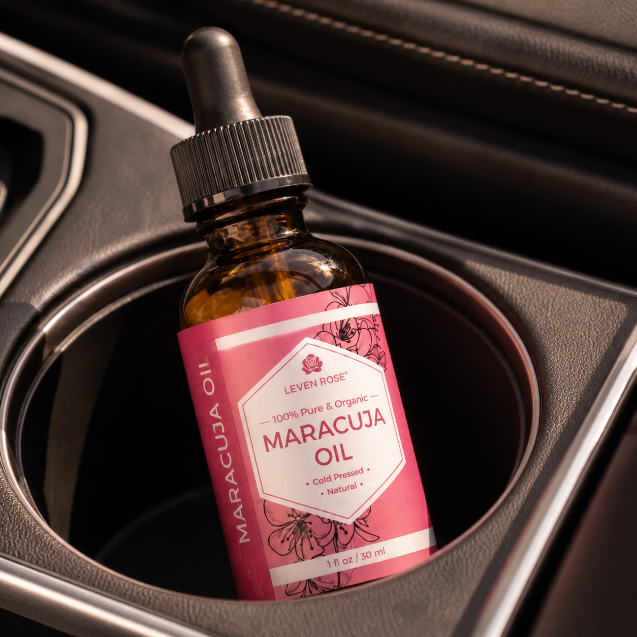 A small amber glass dropper bottle labeled 'Leven Rose Maracuja Oil' resting in a car cup holder. The label is pink and white with floral illustrations, and the bottle has a black dropper cap.
