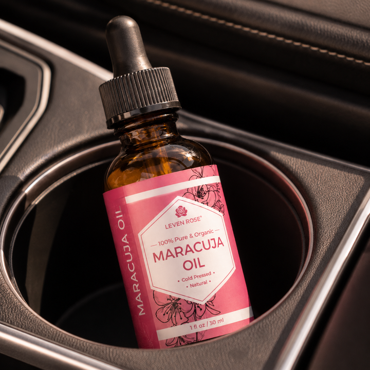A small amber glass dropper bottle labeled 'Leven Rose Maracuja Oil' resting in a car cup holder. The label is pink and white with floral illustrations, and the bottle has a black dropper cap.