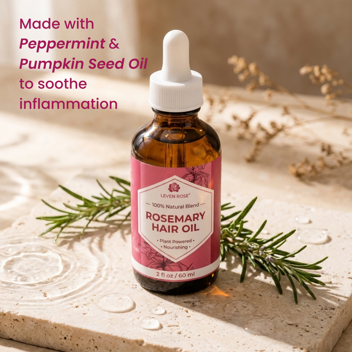 A bottle of Leven Rosemary Hair Oil with a white dropper cap and pink label, placed on a stone surface with rosemary sprigs. Text on the left reads, 'Made with Peppermint & Pumpkin Seed Oil to soothe inflammation.