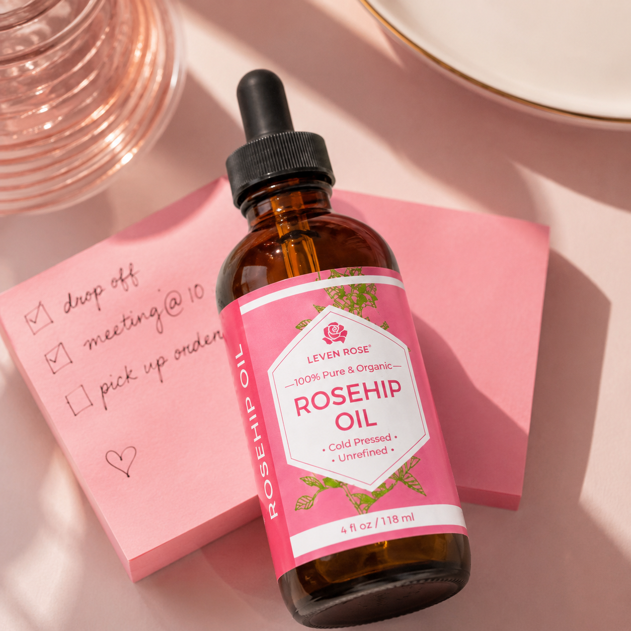 A bottle of Leven Rose Rosehip Oil with a pink label and black dropper cap, styled on a pink background with a pink sticky note and soft-focus pink glassware.