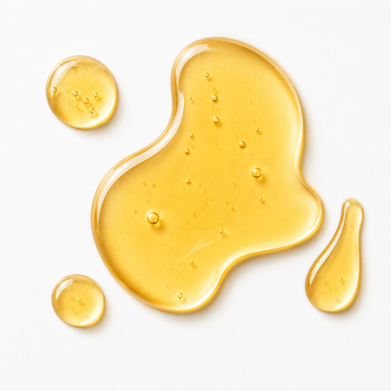 A glossy, translucent golden-yellow liquid, likely an oil or serum, spread across a clean white background in an irregular puddle with three smaller droplets.