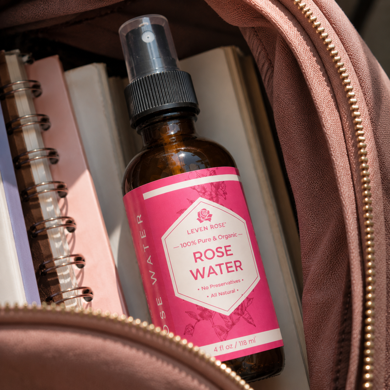 A bottle of Leven Rose Rose Water spray nestled inside a dusty rose zippered pouch, surrounded by notebooks and books.