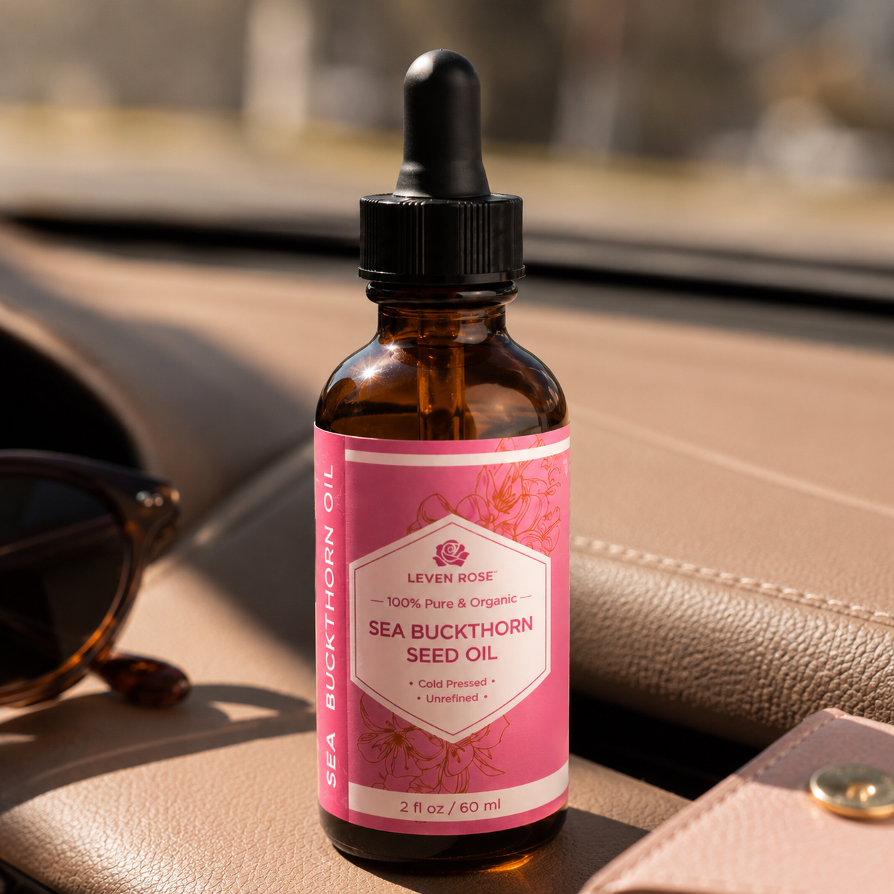 A bottle of Lever Rose Sea Buckthorn Seed Oil with a pink label and black dropper cap, resting on a beige leather surface with sunglasses and a wallet in the background.