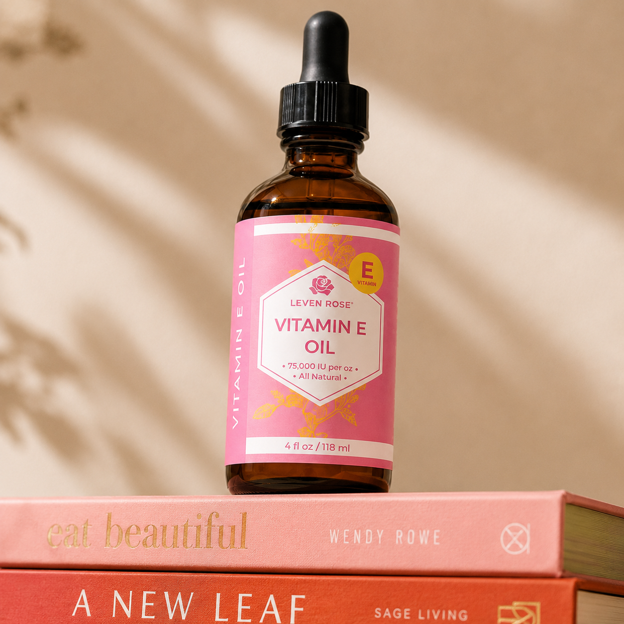 A 4 fl oz amber glass dropper bottle of Leven Rose Vitamin E Oil with a pink label, resting on top of two stacked pink books against a warm beige background with soft leaf shadows.