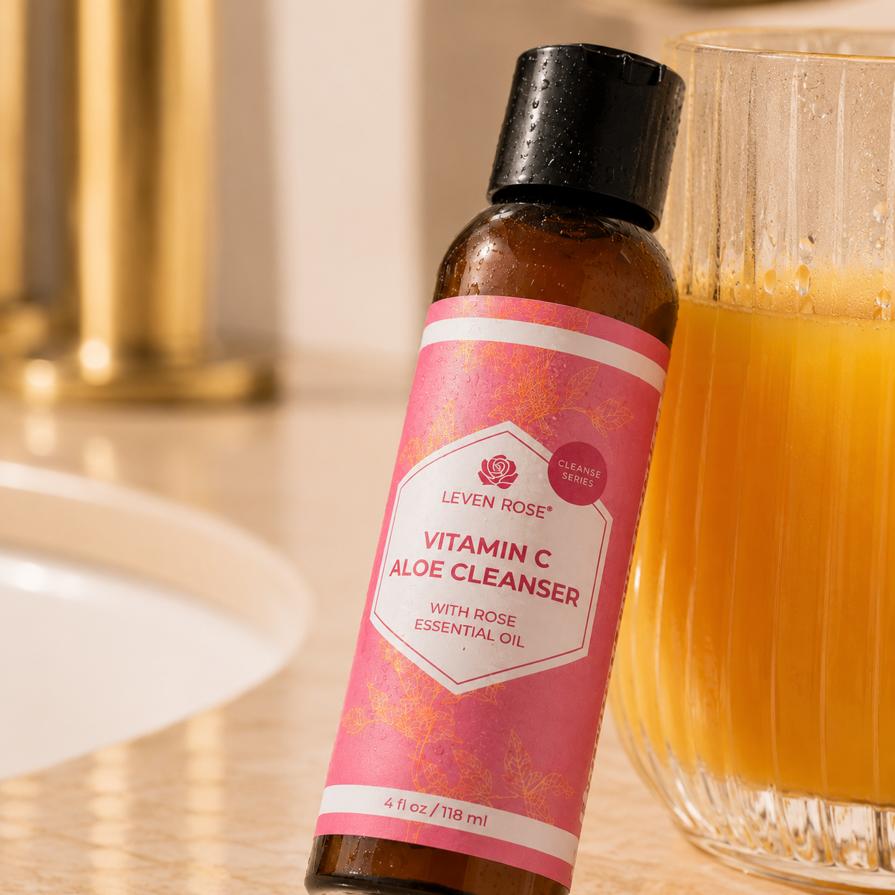 A bottle of Leven Rose Vitamin C Aloe Cleanser with a pink label and black cap, shown on a bathroom counter next to a glass of orange juice.