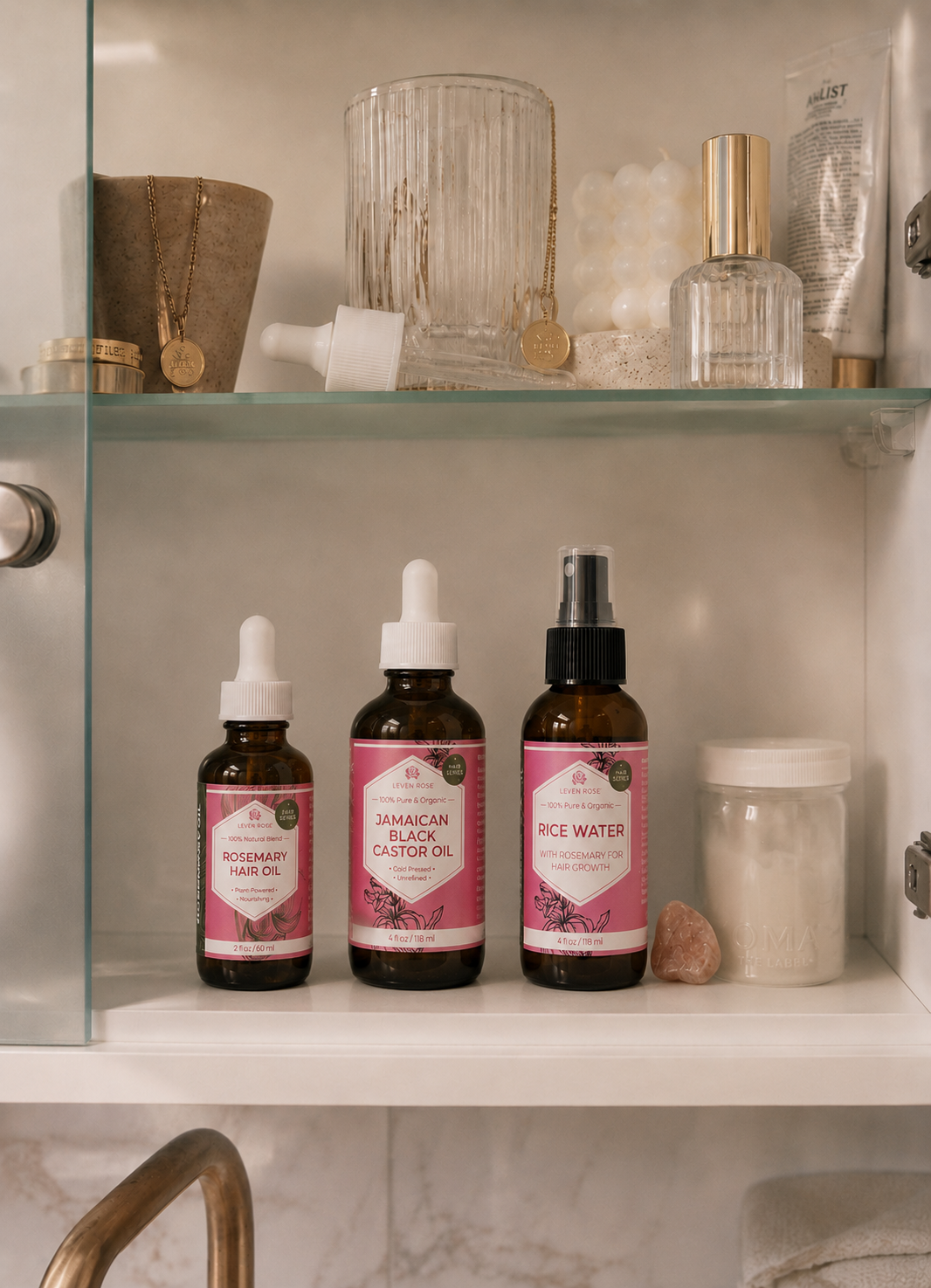 Three hair care bottles from The Art of Hair on a bathroom shelf: Rosemary Hair Oil, Swayvan Black Cucumber Oil, and Rice Water. The bottles are amber glass with pink labels and white dropper and spray caps, displayed next to a clear jar.