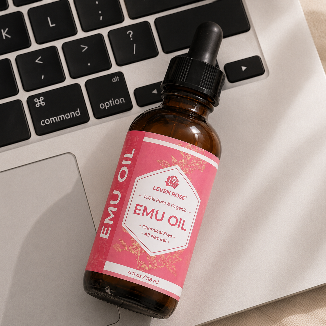 A bottle of Leven Rose EMU OIL with a pink label and black dropper cap, resting on a silver laptop keyboard.