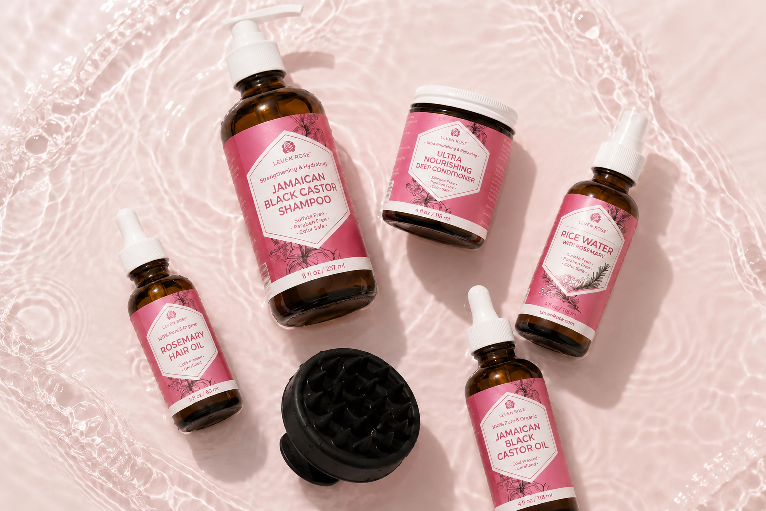Six haircare products from The Art of Blackness are arranged on a pink textured surface with a white lace-like pattern. The products include shampoo, conditioner, honey water, rosemary hair oil, Jamaican black castor shampoo, and a black scalp massager.