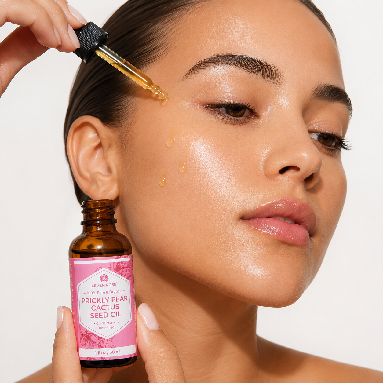 A close-up of a woman applying seed oil from a dropper to her face while holding a small amber bottle labeled 'Let's Be Rose Prickly Pear Cactus Seed Oil' against a clean white background.
