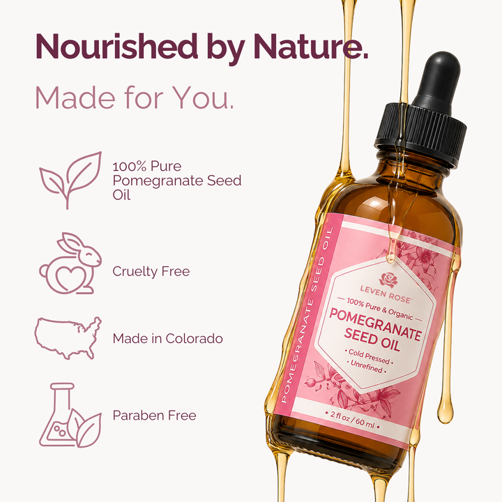 A bottle of Leven Rose Pomegranate Seed Oil with a pink label and black dropper cap, shown on a light background with decorative text and icons about pure pomegranate seed oil, cruelty free, made in Colorado, and paraben free. Gold-colored oil is dripping around the bottle.